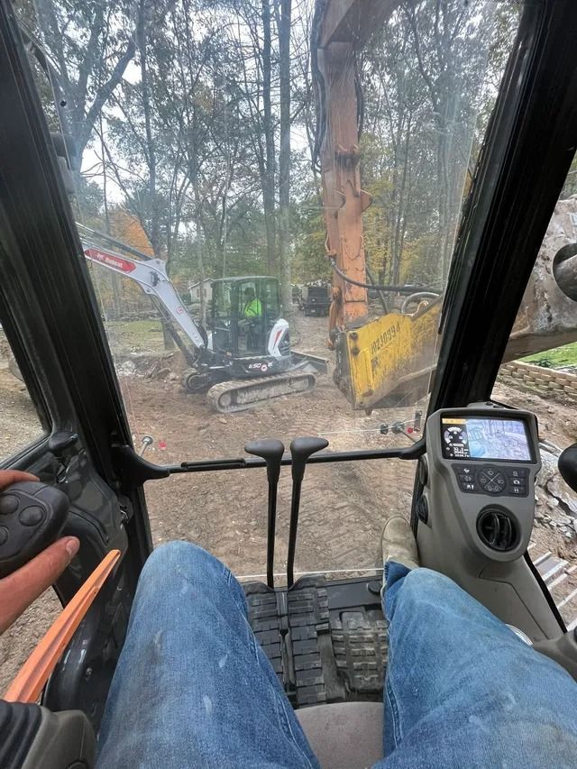 Inside an excavator cab, a worker operates controls while clearing a wooded site with another machine outside.