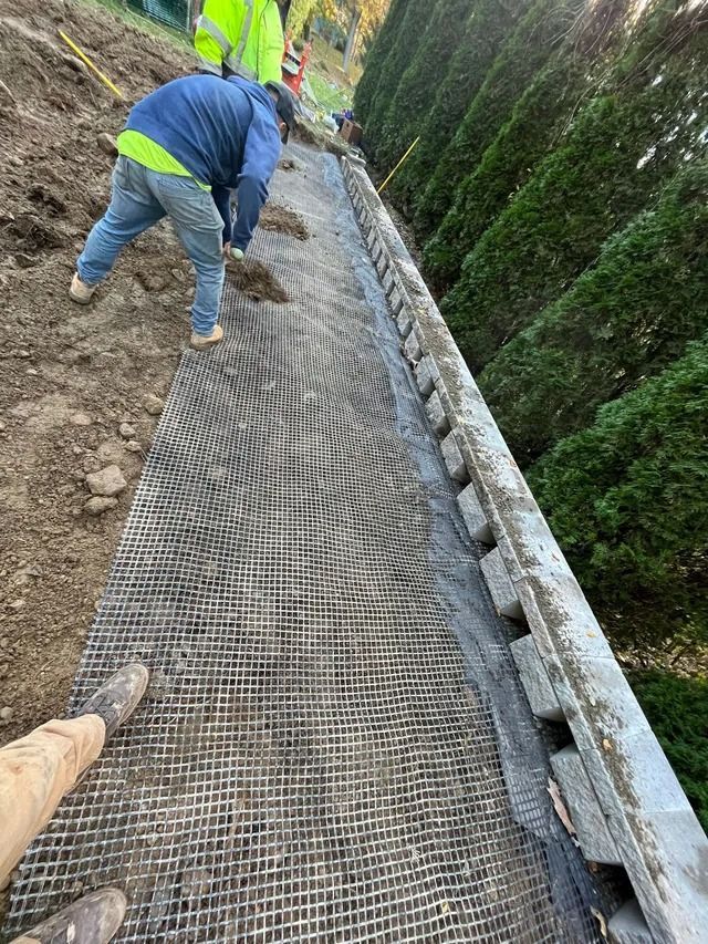 Workers installing metal mesh on a steep outdoor path beside a hedge and retaining wall