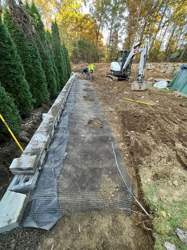 Construction site with excavator and workers beside a long trench lined with black mesh and stones