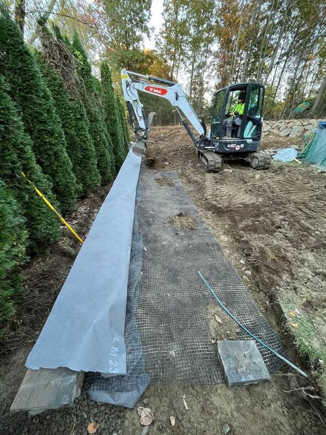 Excavator leveling soil beside a long gray edging strip along a hedge in a backyard.