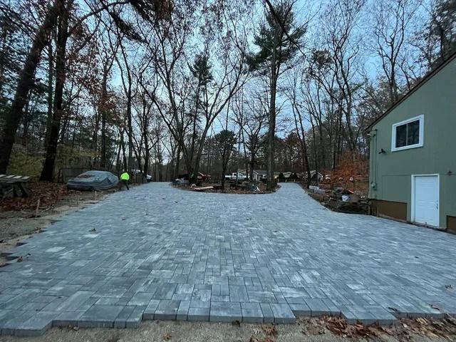 Wide gray paver driveway beside a beige house, bordered by leafless trees in a wooded yard