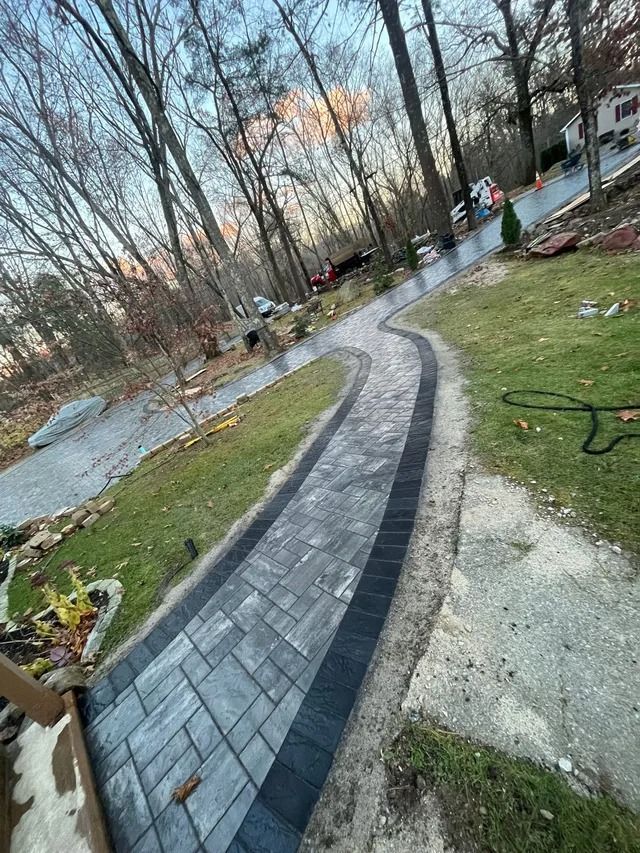 Curved brick walkway through a leafless yard with grass, gravel edges, and a cloudy sunset sky