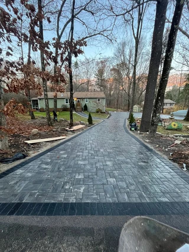 Paved driveway leading to a house in a wooded yard with bare trees and garden beds