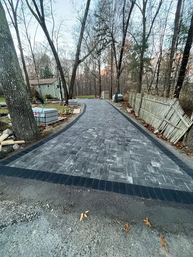 Paved driveway or walkway under construction, lined with bare trees and a wooden fence