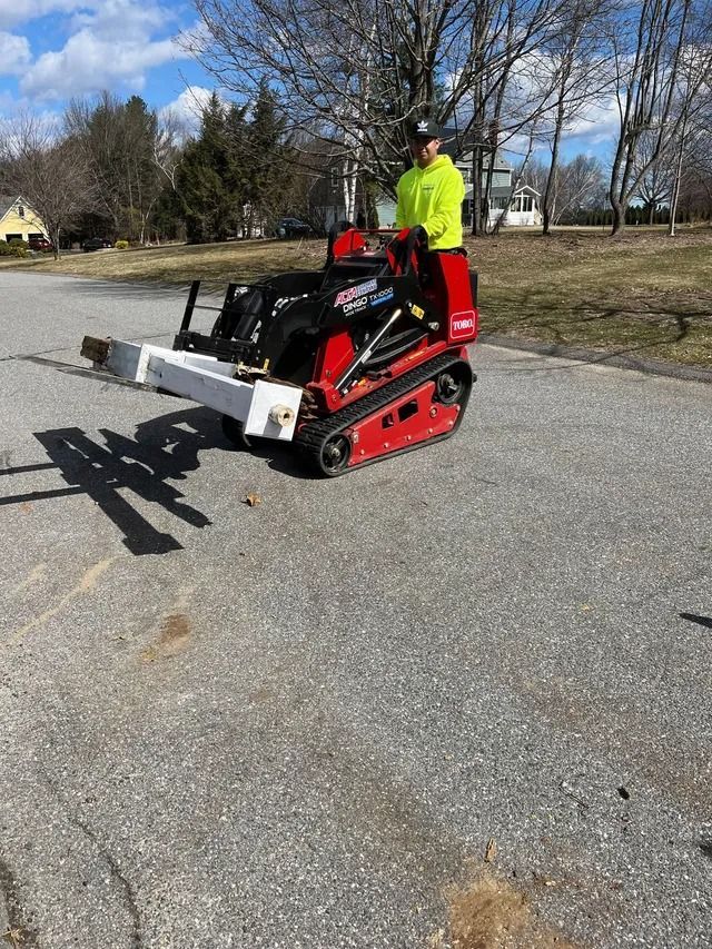 Person standing on a red tracked mini loader in a driveway, holding a wide white attachment