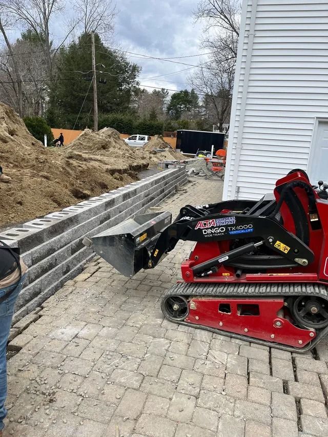 Red tracked mini skid steer parked beside a stacked retaining wall on a paved side yard construction site.