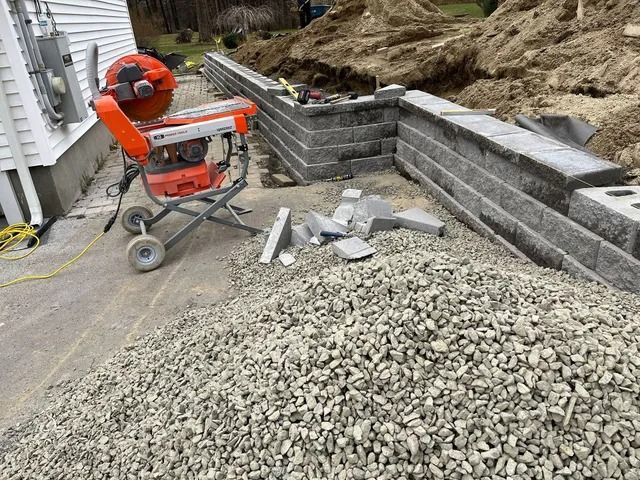 Construction site with orange masonry saw, gravel pile, and newly built gray block retaining wall