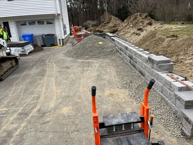 Driveway construction site with gravel, retaining wall blocks, and equipment near a house.