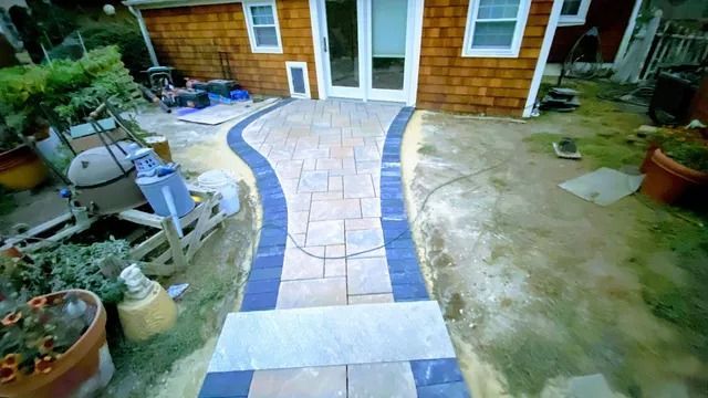 Curved stone walkway through a yard leading to a house, with patio items and plants on both sides