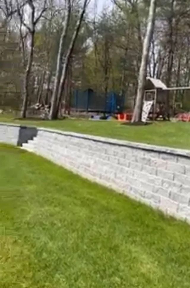 Backyard with a raised stone retaining wall, green lawn, and trees around a small play area