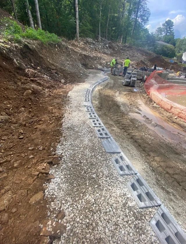 Construction crew installing drainage along a muddy hillside road beside orange safety fencing