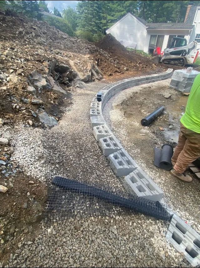 Construction site with gravel path, cinder blocks, black drain pipe, and worker in neon shirt beside a house