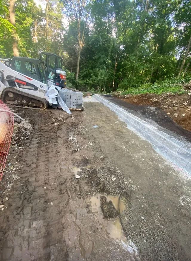 Small excavator on muddy roadside beside a fresh concrete drainage channel in a wooded area