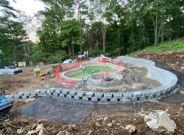 Construction site with a curved stone retaining wall and workers near a partially built circular structure in a wooded area