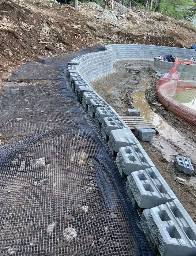 Curved gray block retaining wall under construction beside a dirt slope and black mesh on a hillside.