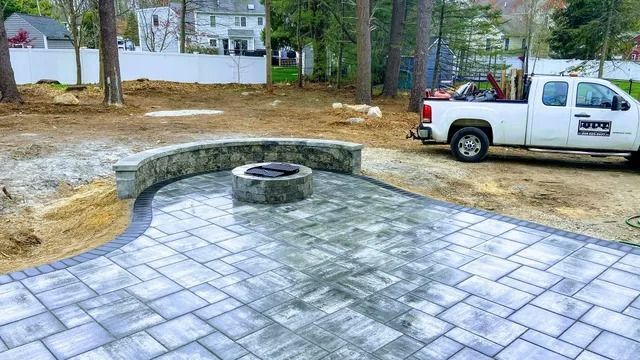 Patio pavers with a curved stone fire pit beside a white pickup truck in a wooded yard