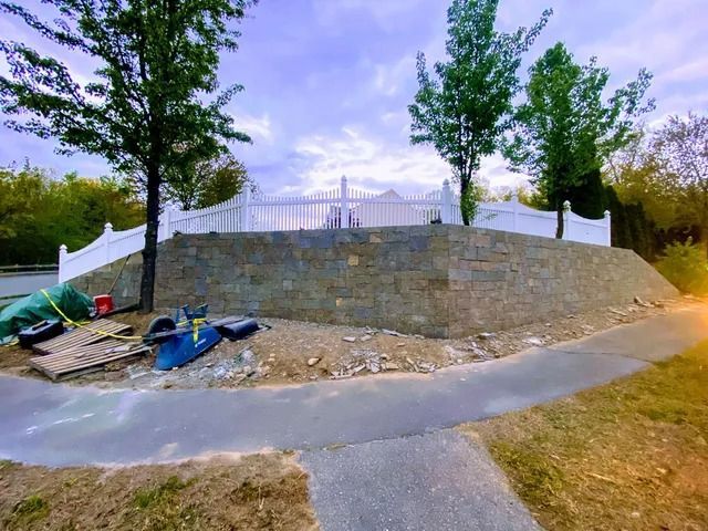 Stone retaining wall along a curved path with trees and fencing in a park-like area