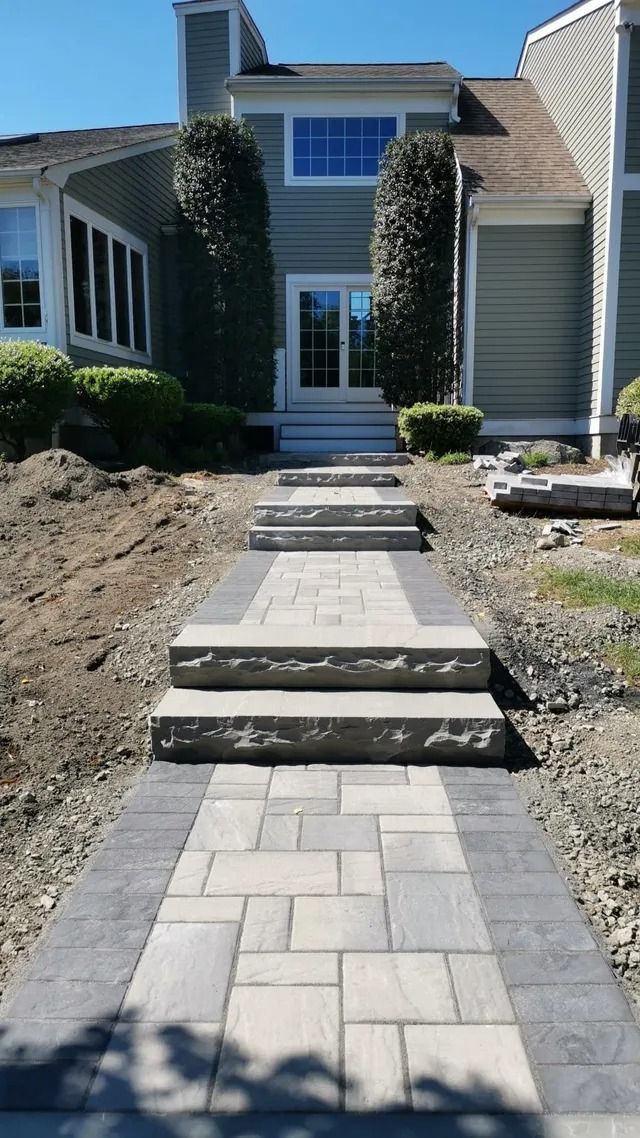 Front walkway to a house with concrete steps and narrow stone path, bordered by dirt landscaping.