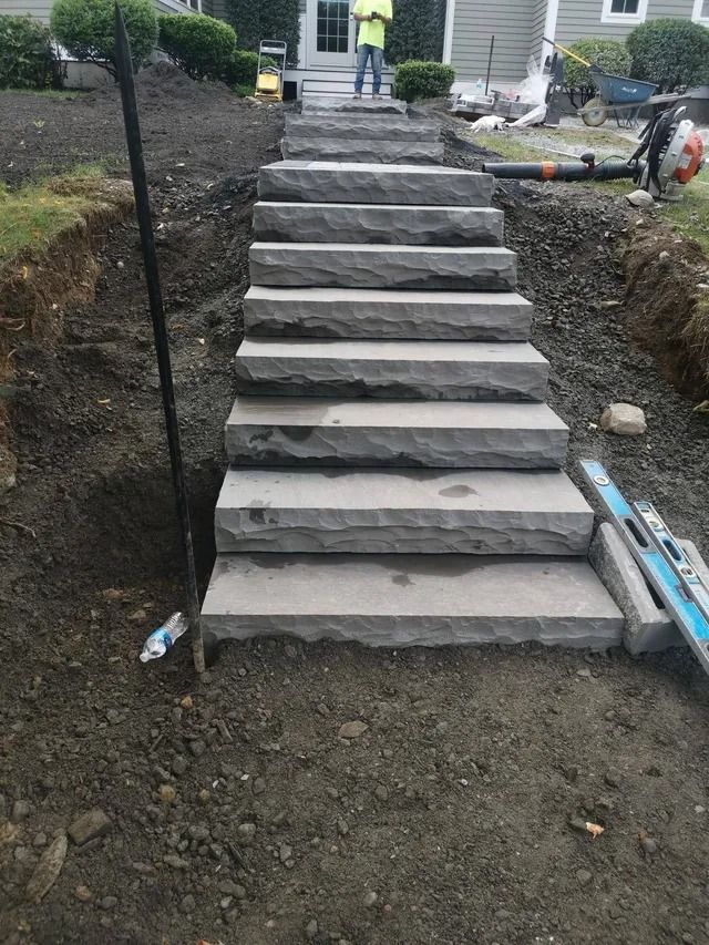 Stone steps under construction in a dirt slope, with a person standing at the top and tools nearby