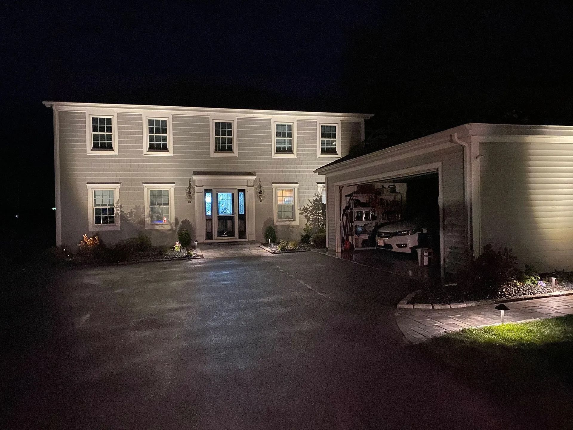 Two-story house at night with porch lights on and an open garage beside a dark driveway.