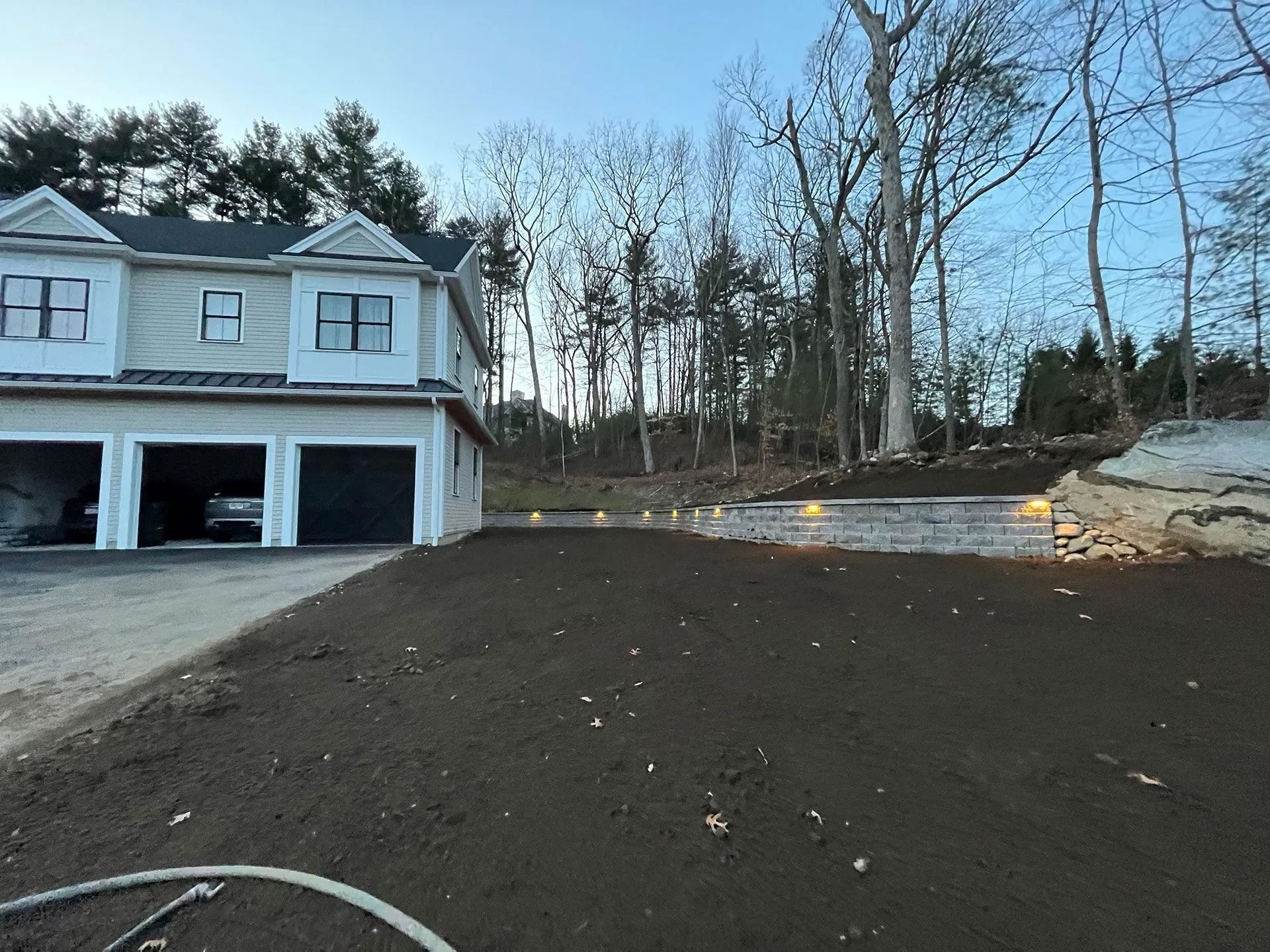 New house with two-car garage beside a freshly graded dirt lot and wooded hillside at dusk