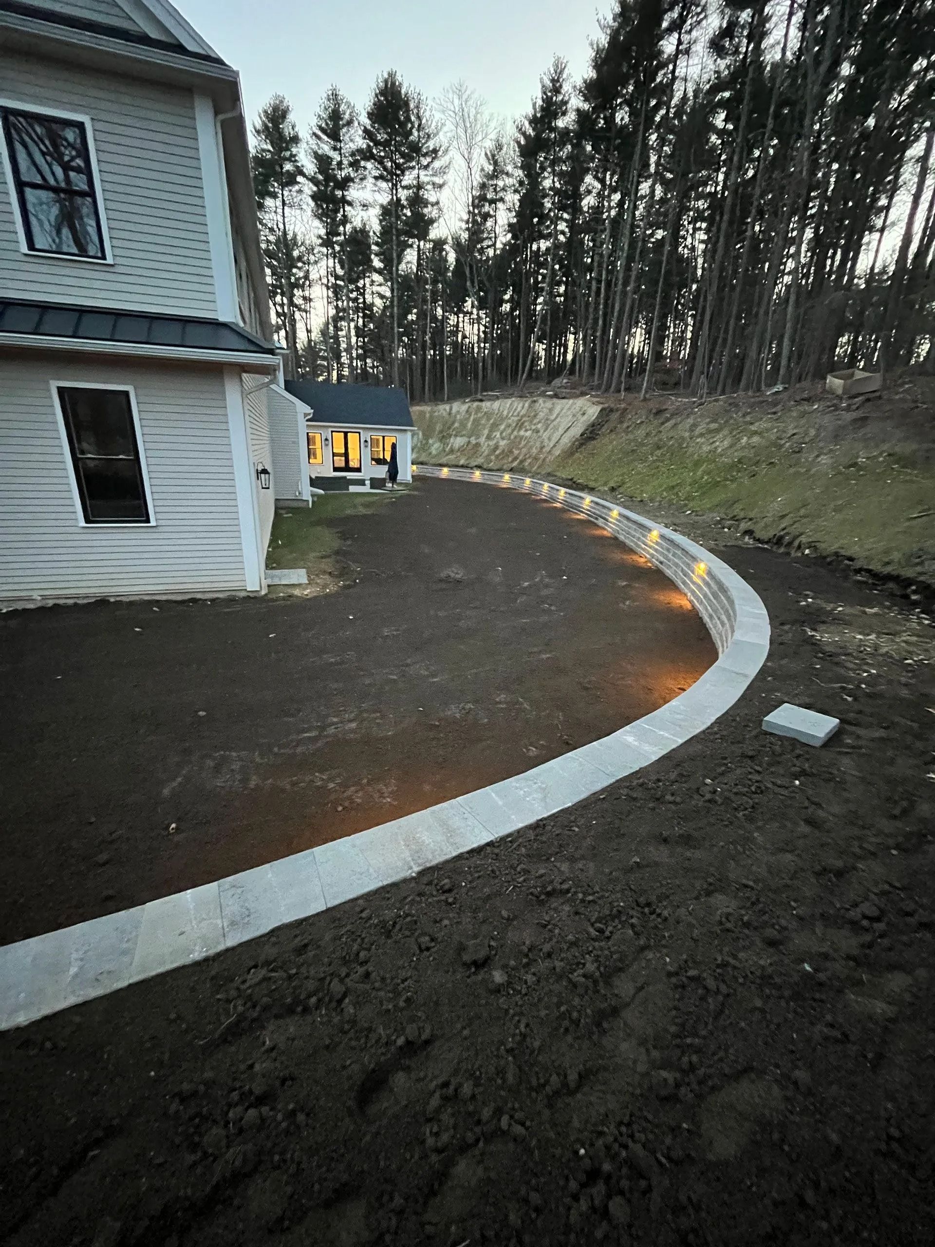 Curved concrete walkway beside a white house, leading toward a small yellow outbuilding at dusk.