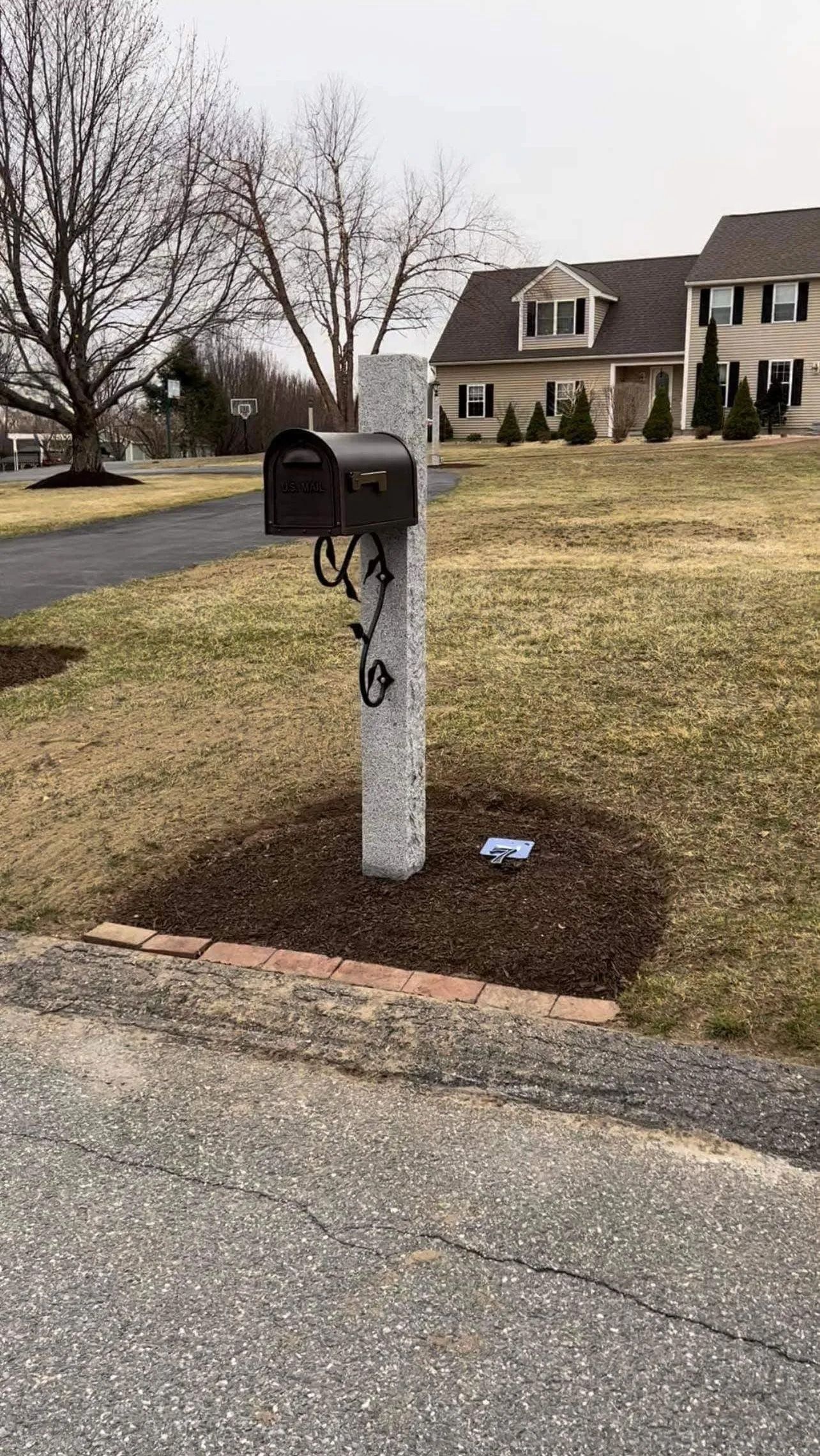 Mailbox on a white post beside a suburban driveway, with a house and bare trees in the background.