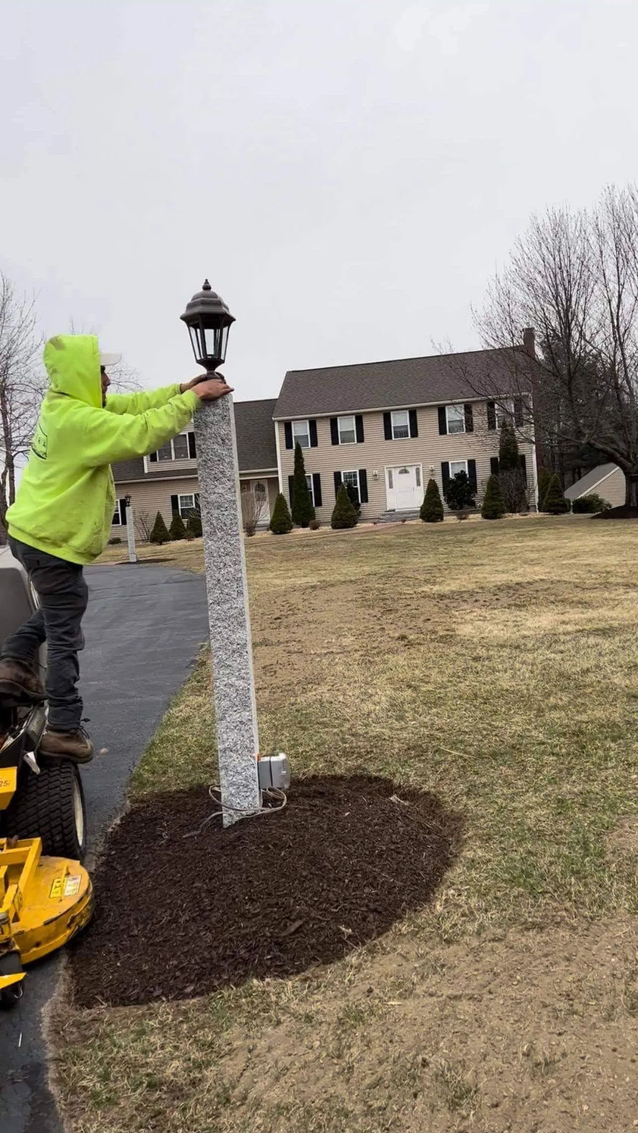 Person in a neon jacket installing a lamp post beside a sidewalk in a suburban yard.