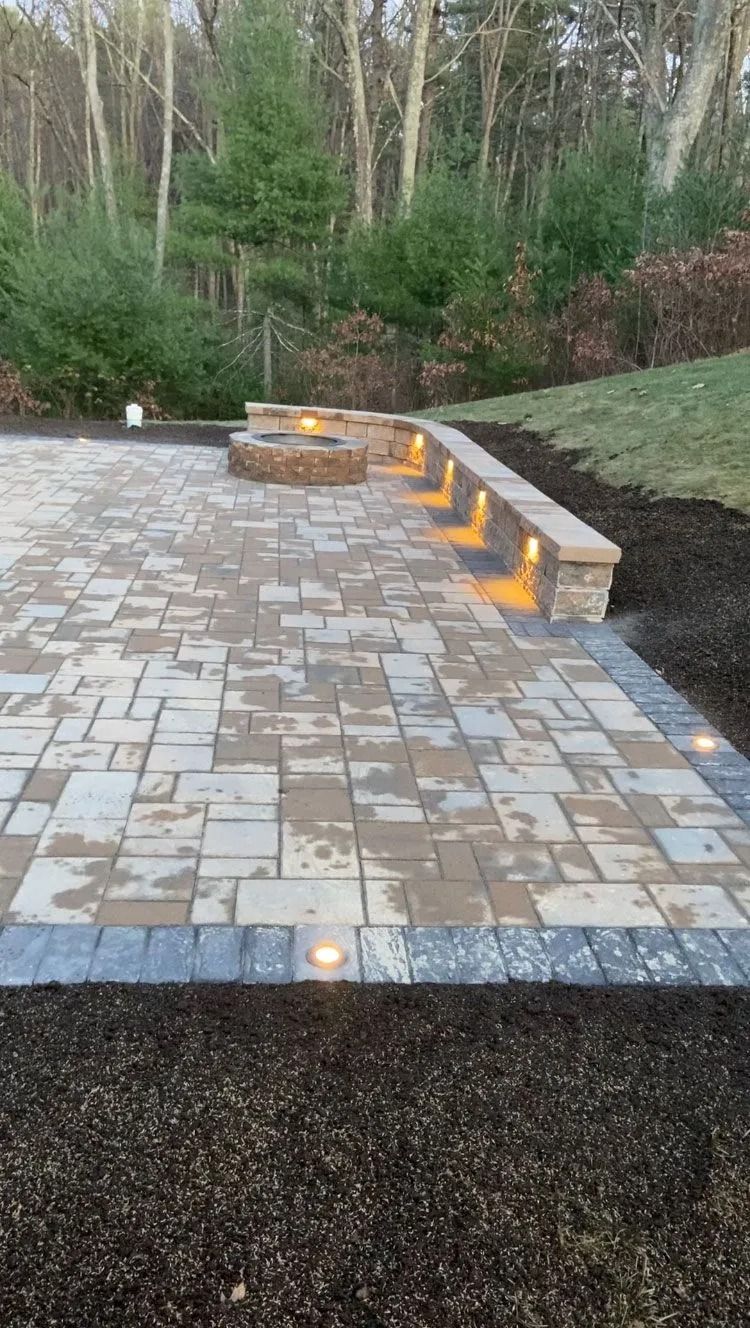 Paved patio with curved stone steps and yellow lights, bordered by mulch and trees at dusk