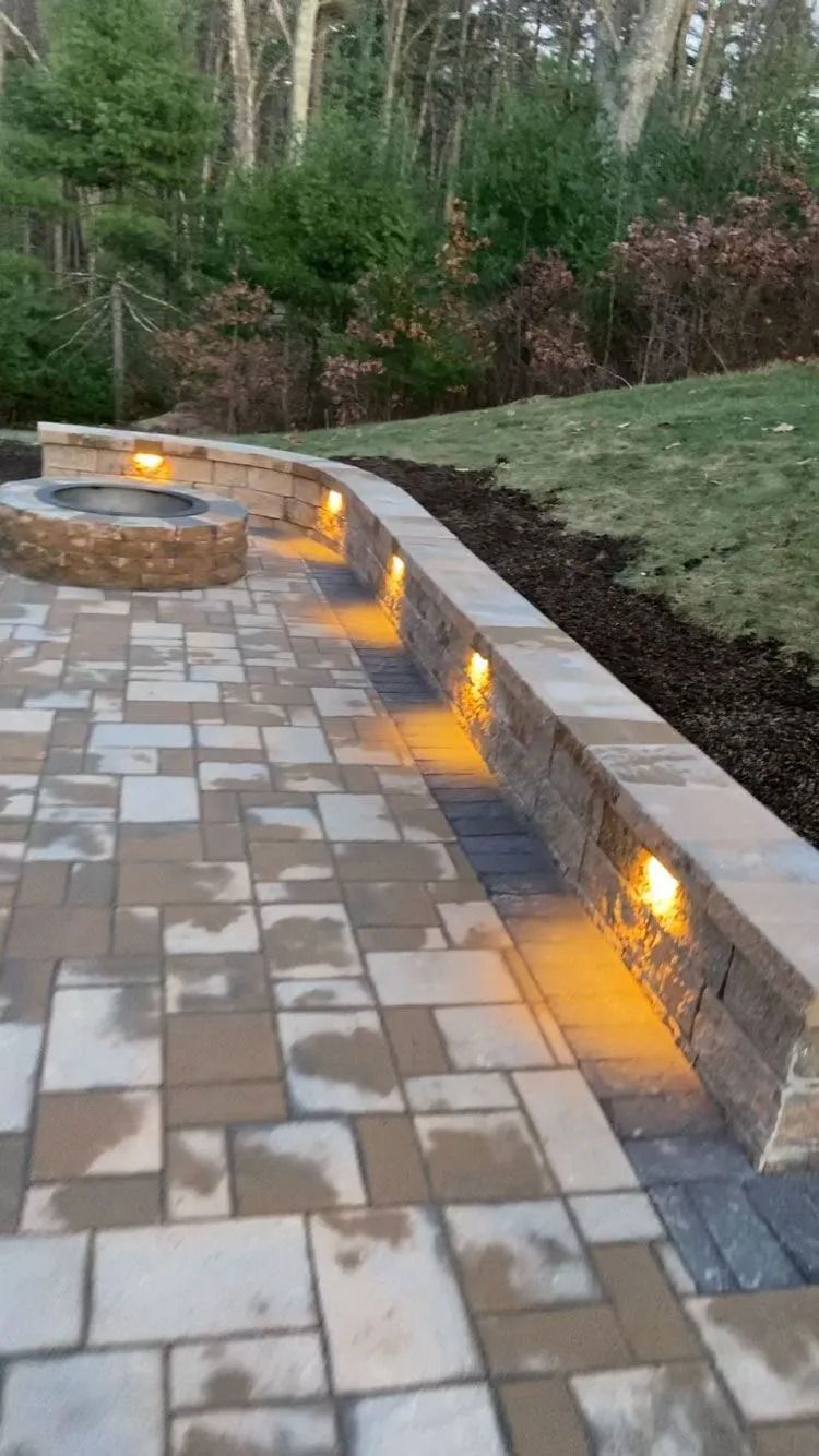 Outdoor stone patio with curved low wall and warm recessed lights at dusk, backed by trees and grass