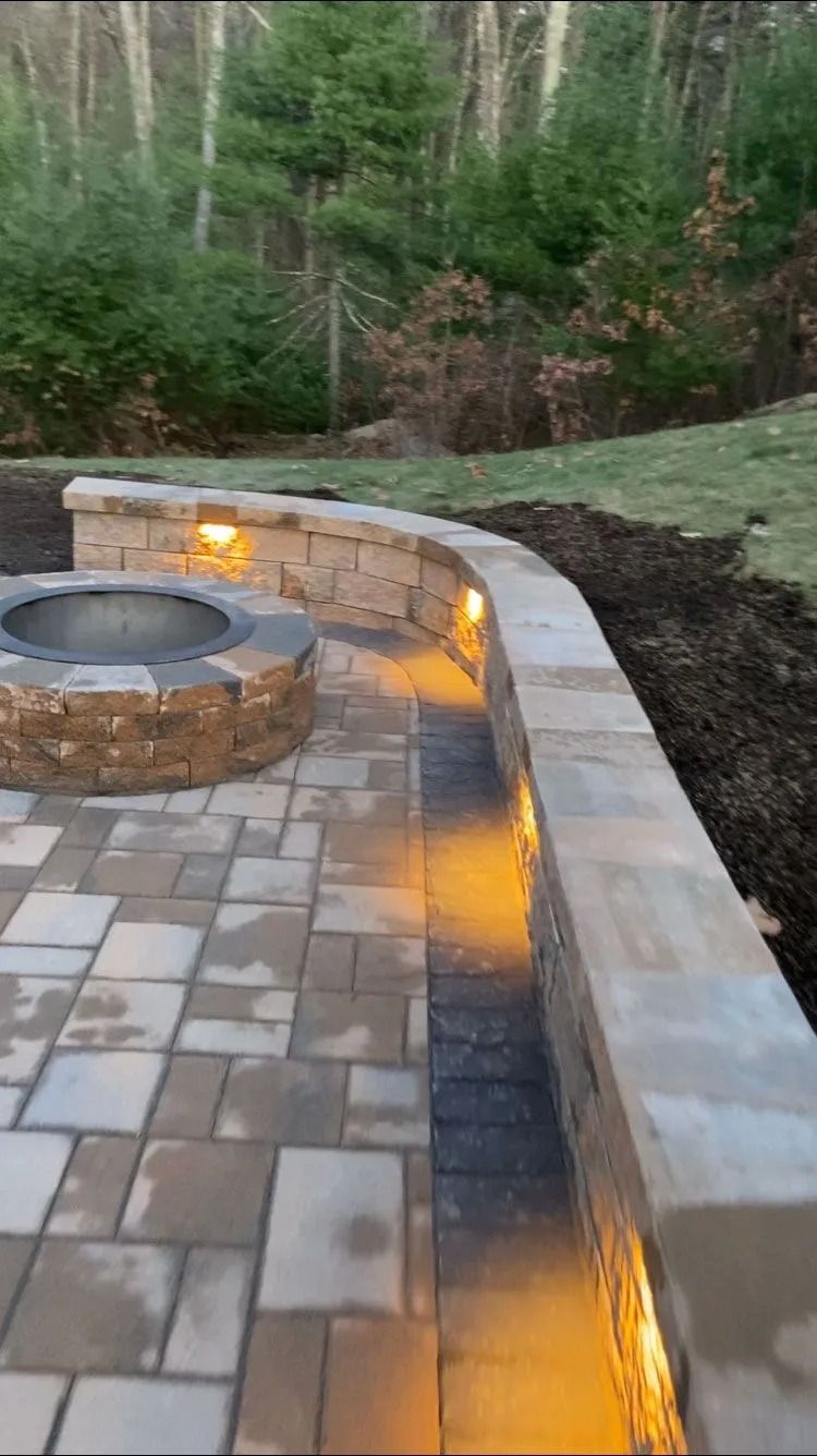 Backyard patio with a curved stone fire pit and warm accent lighting beside a wooded slope