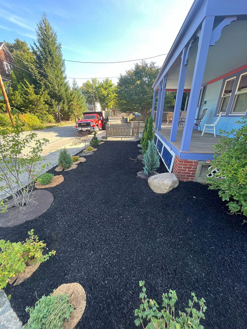 Front yard with black gravel path, red tractor, and blue porch beside trees and shrubs.