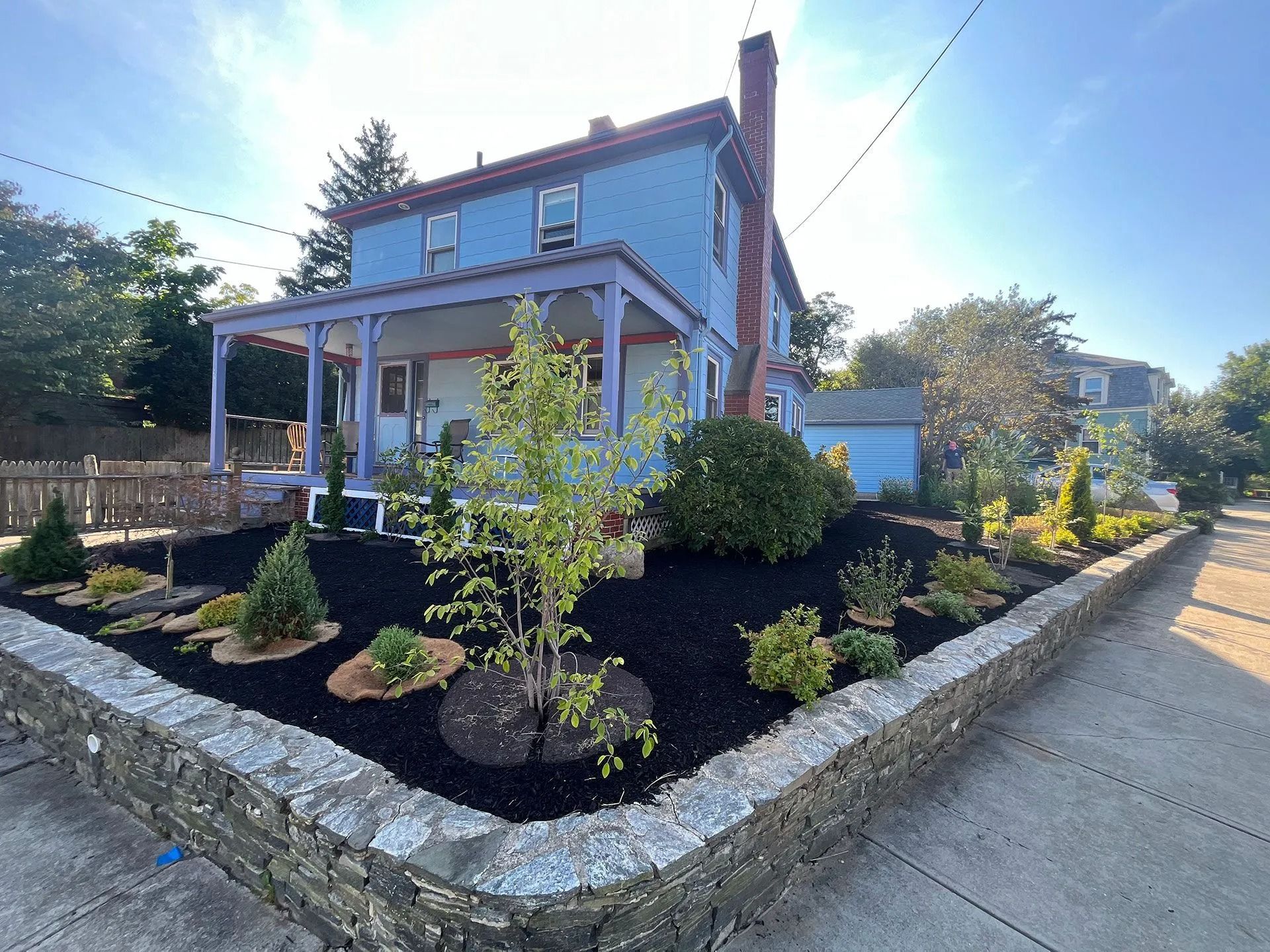 Two-story blue house with a stone border, landscaped front yard, and driveway in bright sunlight