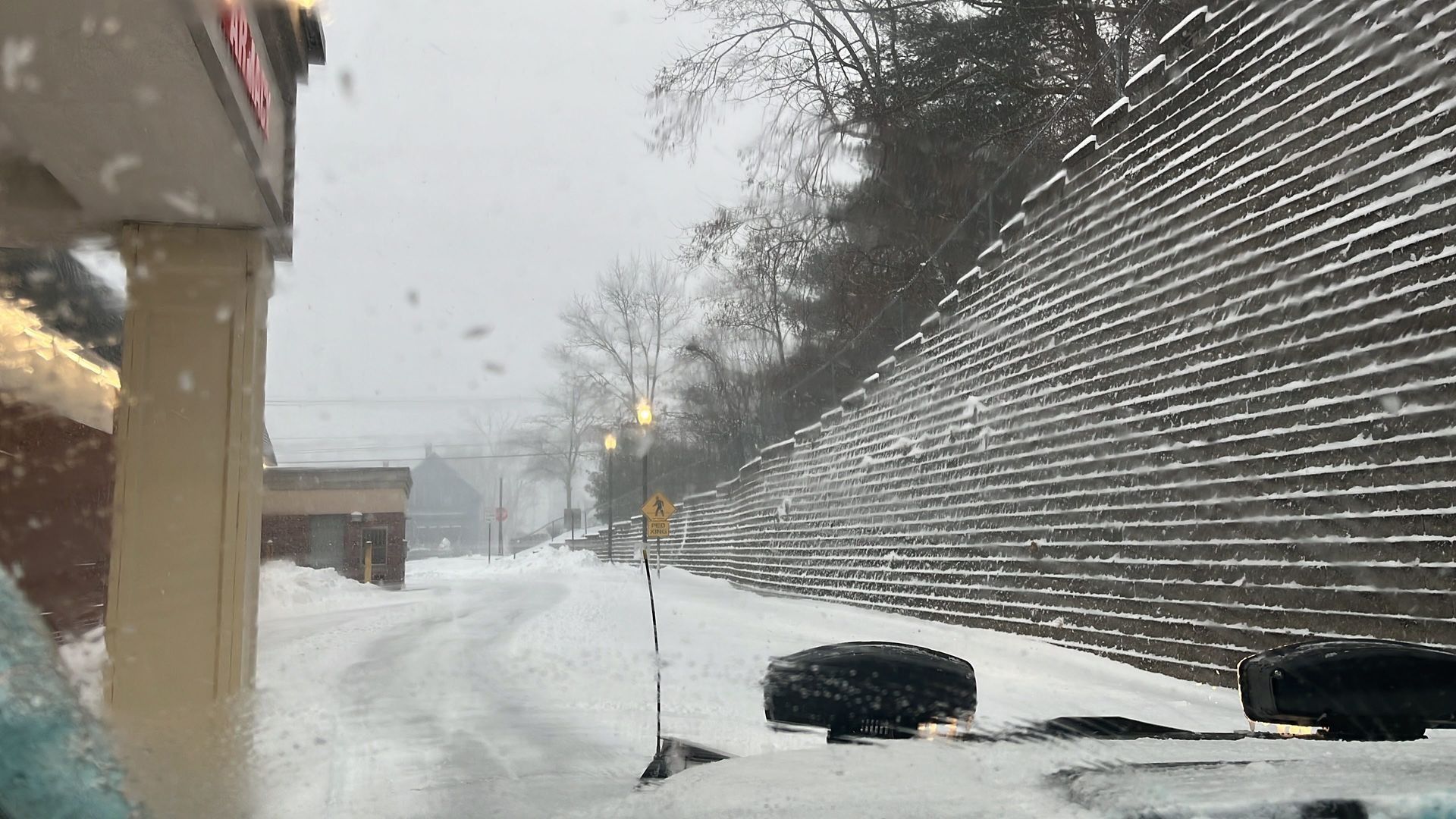 Snowy road beside a building and retaining wall, with heavy snowfall reducing visibility.