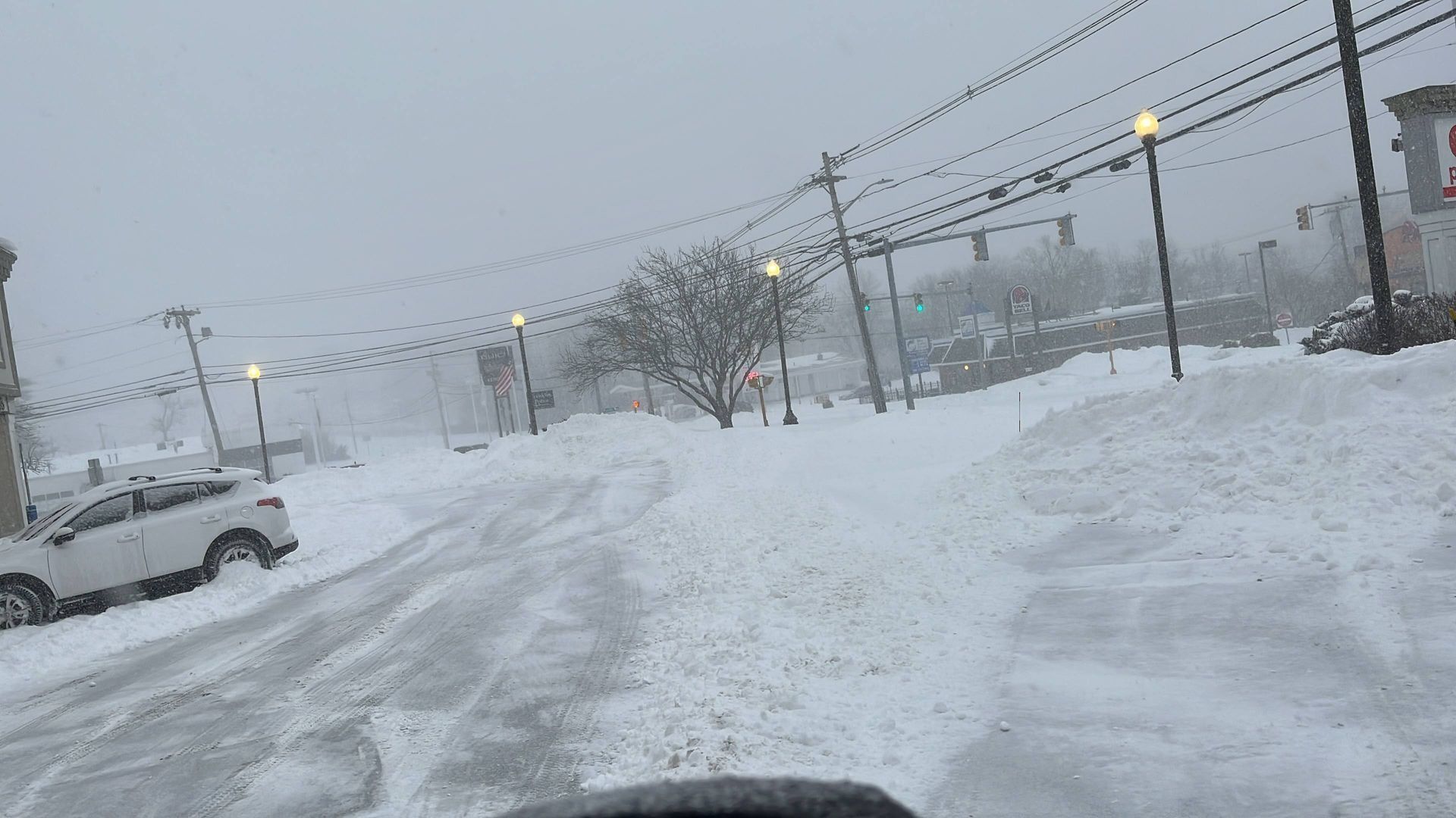 Snow-covered street with parked car, power lines, and glowing streetlights in a heavy snowfall