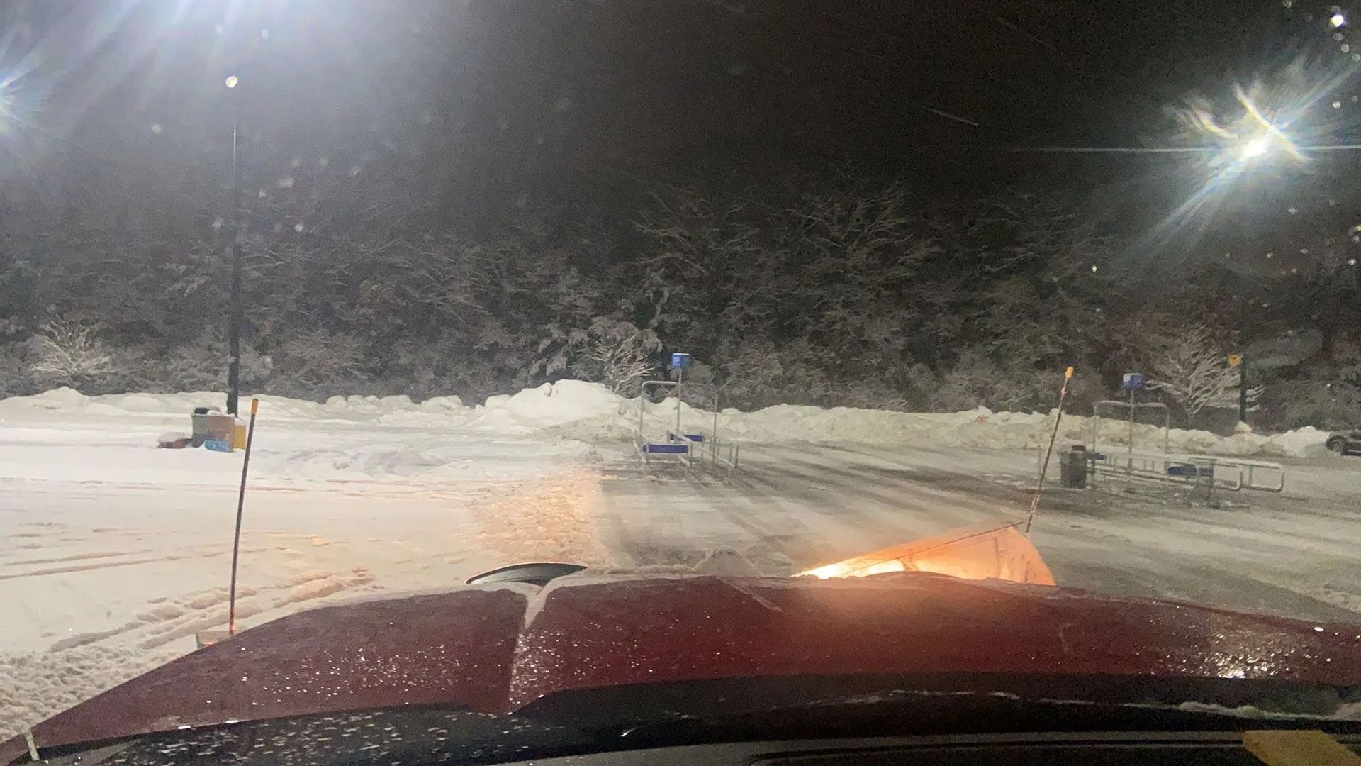 Snowy parking lot seen from inside a truck at night, with headlights and heavy snow on the windshield.