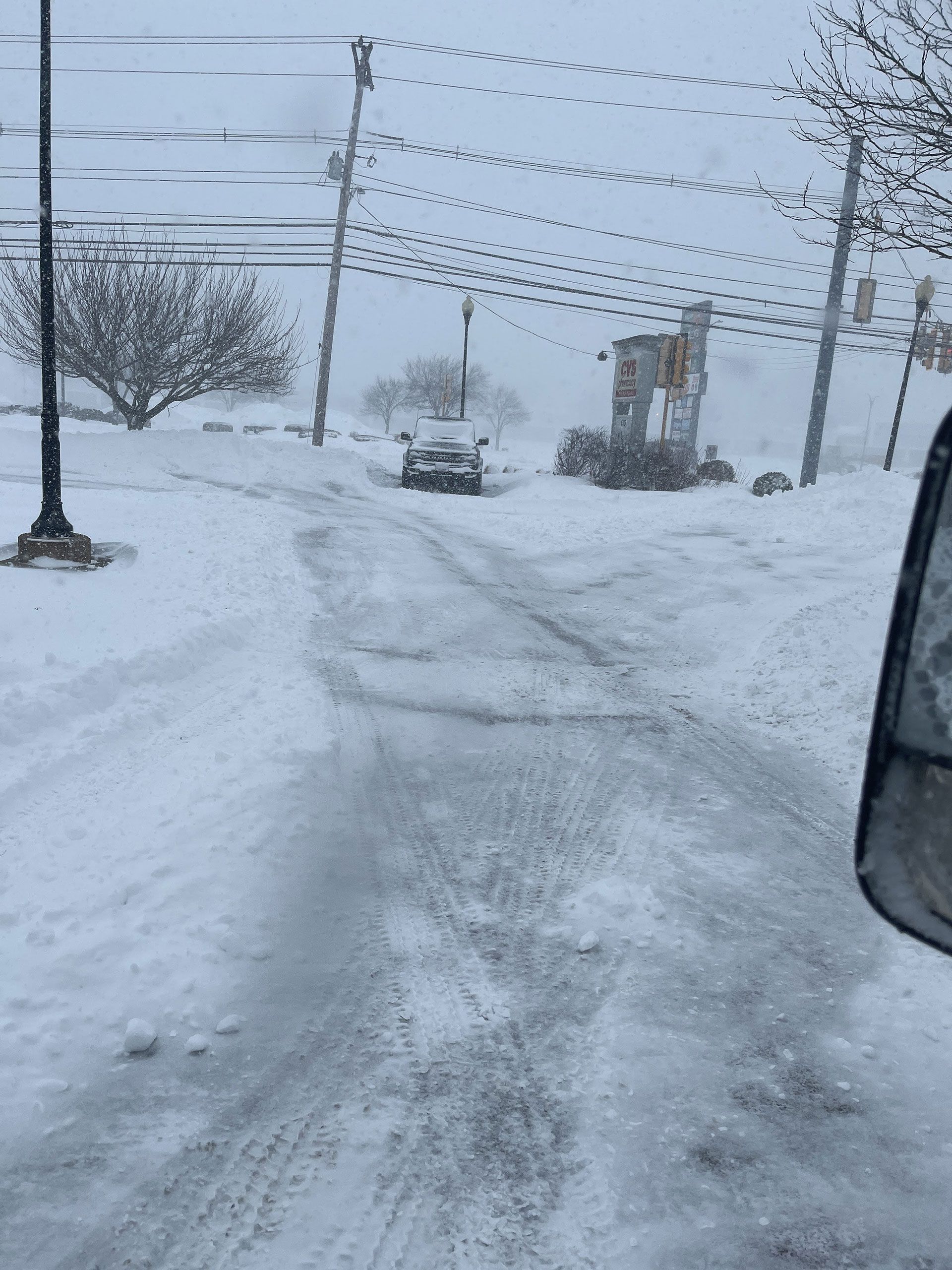 Snow-covered road with tire tracks, power lines, and a parked car in a winter storm