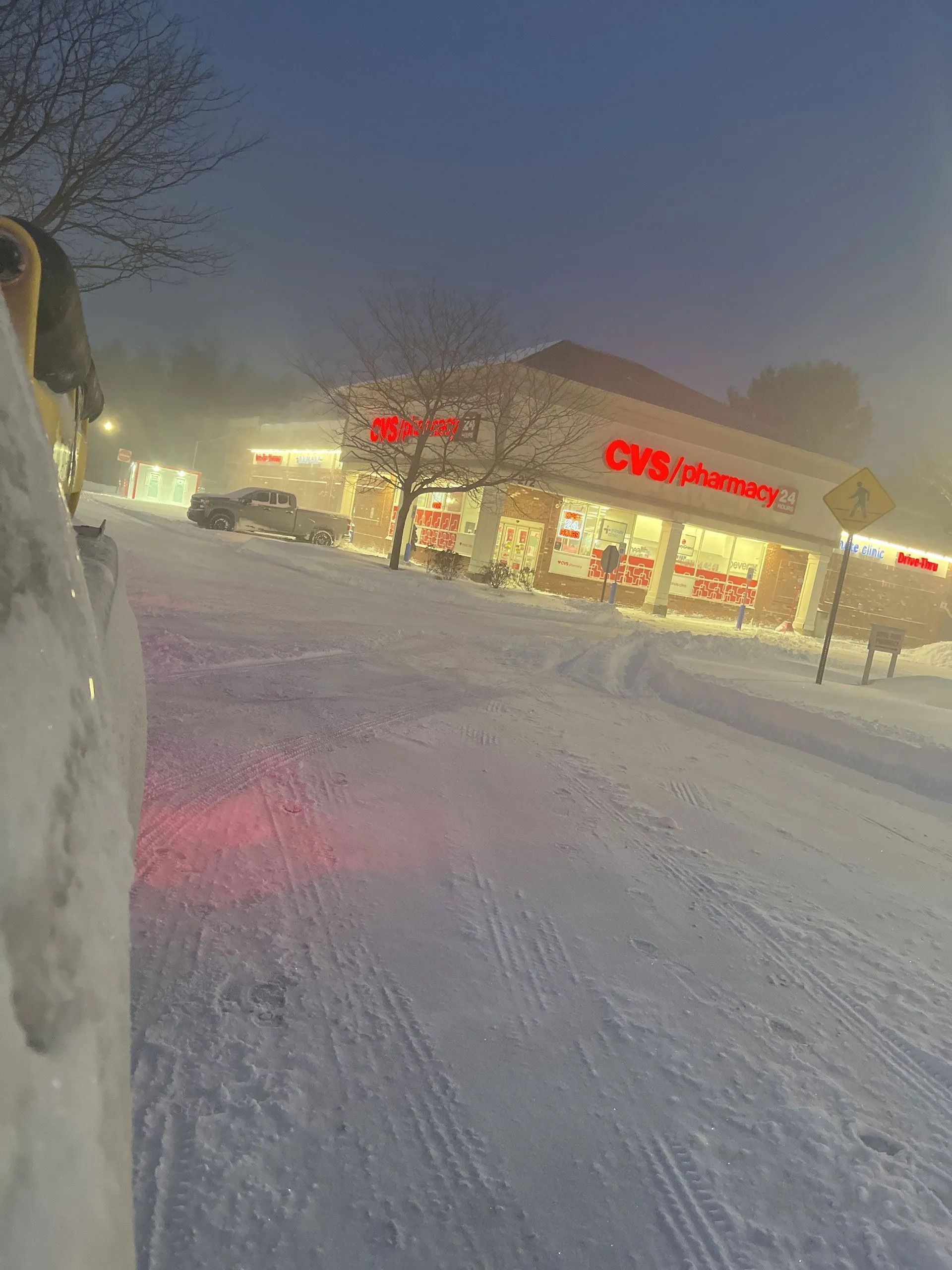 Snowy street at dusk with a lit CVS Pharmacy storefront and heavy snowfall reducing visibility
