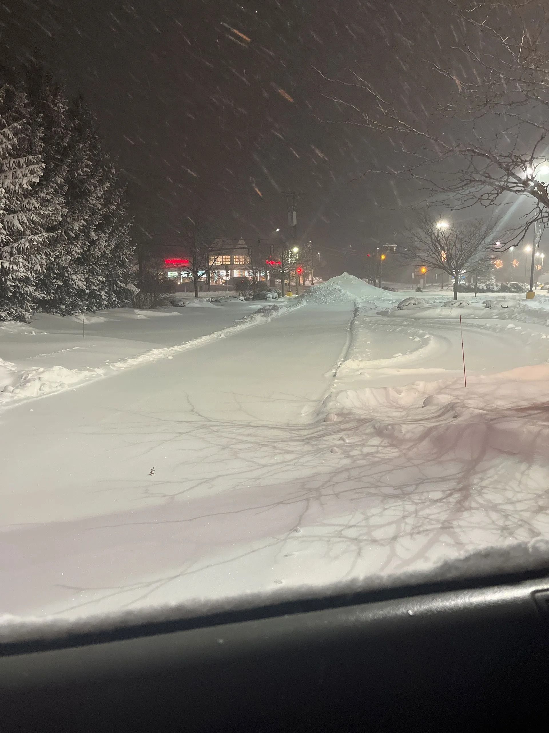 Snowy road at night with heavy snowfall, plowed lanes, and distant glowing storefront signs