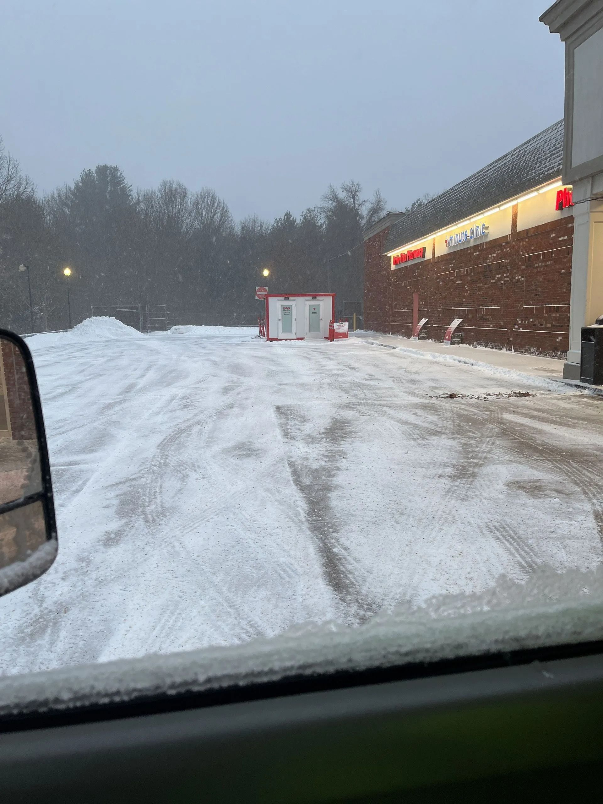 Snowy parking lot beside a brick store at dusk, seen from inside a car.
