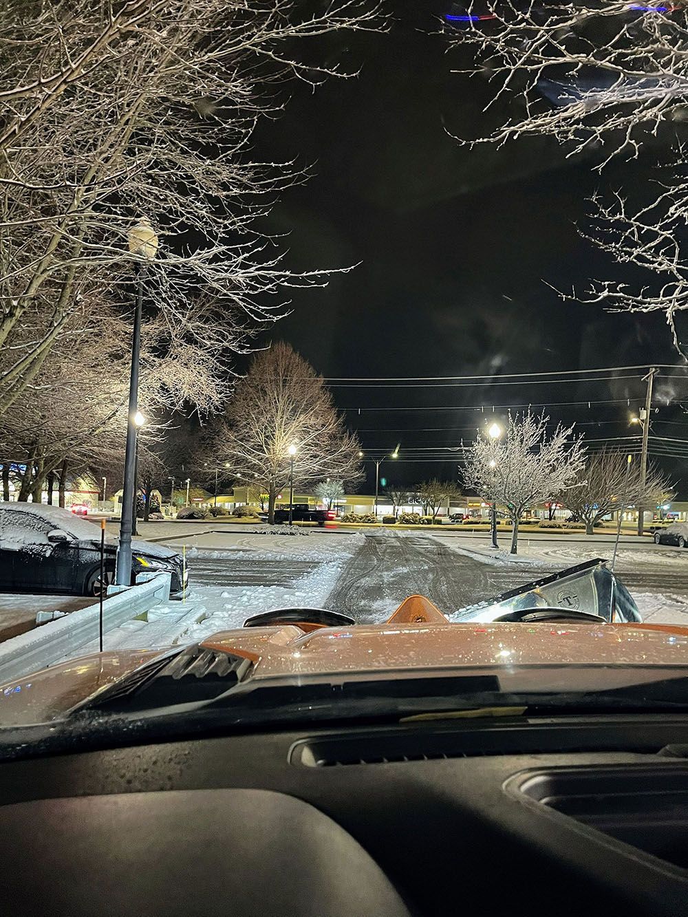 Nighttime view from a car windshield of a snowy parking lot with lit trees and parked cars