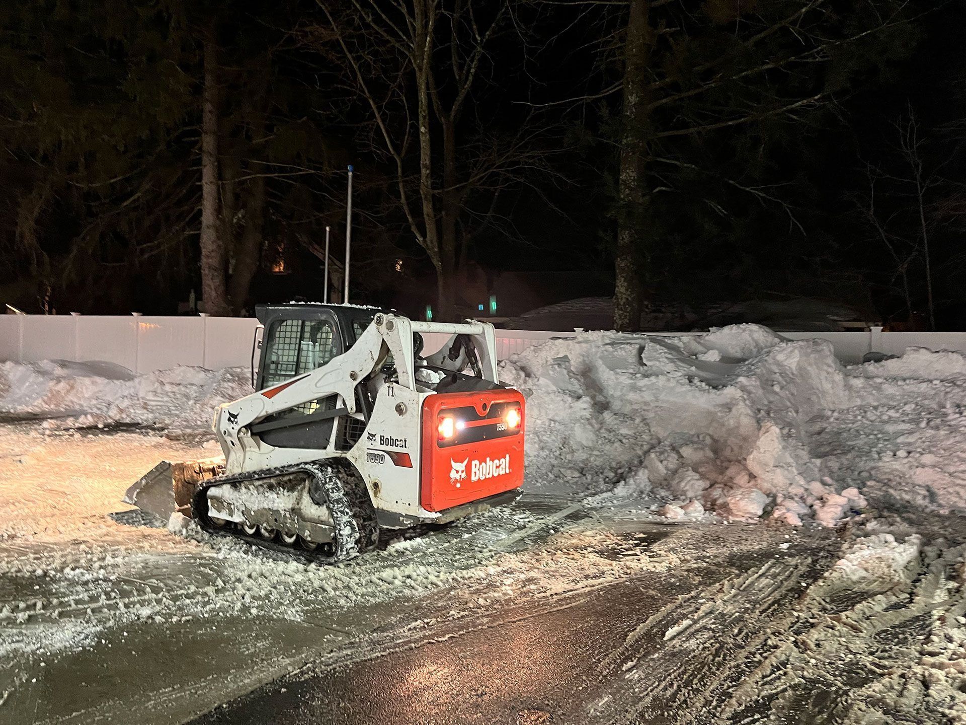 Compact snow loader clearing a snowy roadside at night, with red rear panel and headlights on.