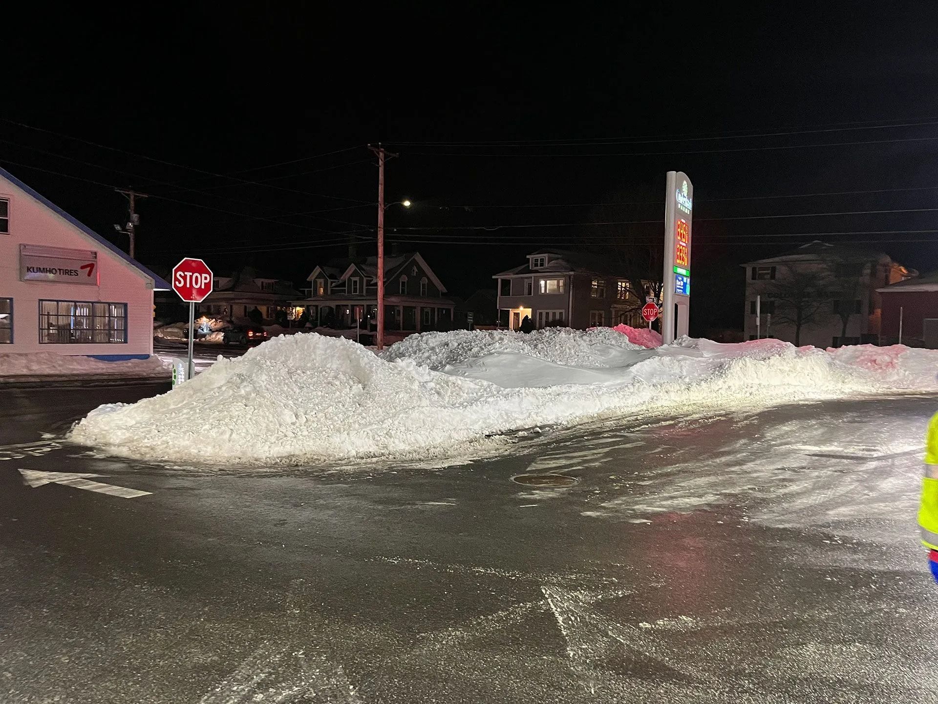 Snow-covered parking lot at night with a large snow pile, stop sign, and illuminated gas station sign.