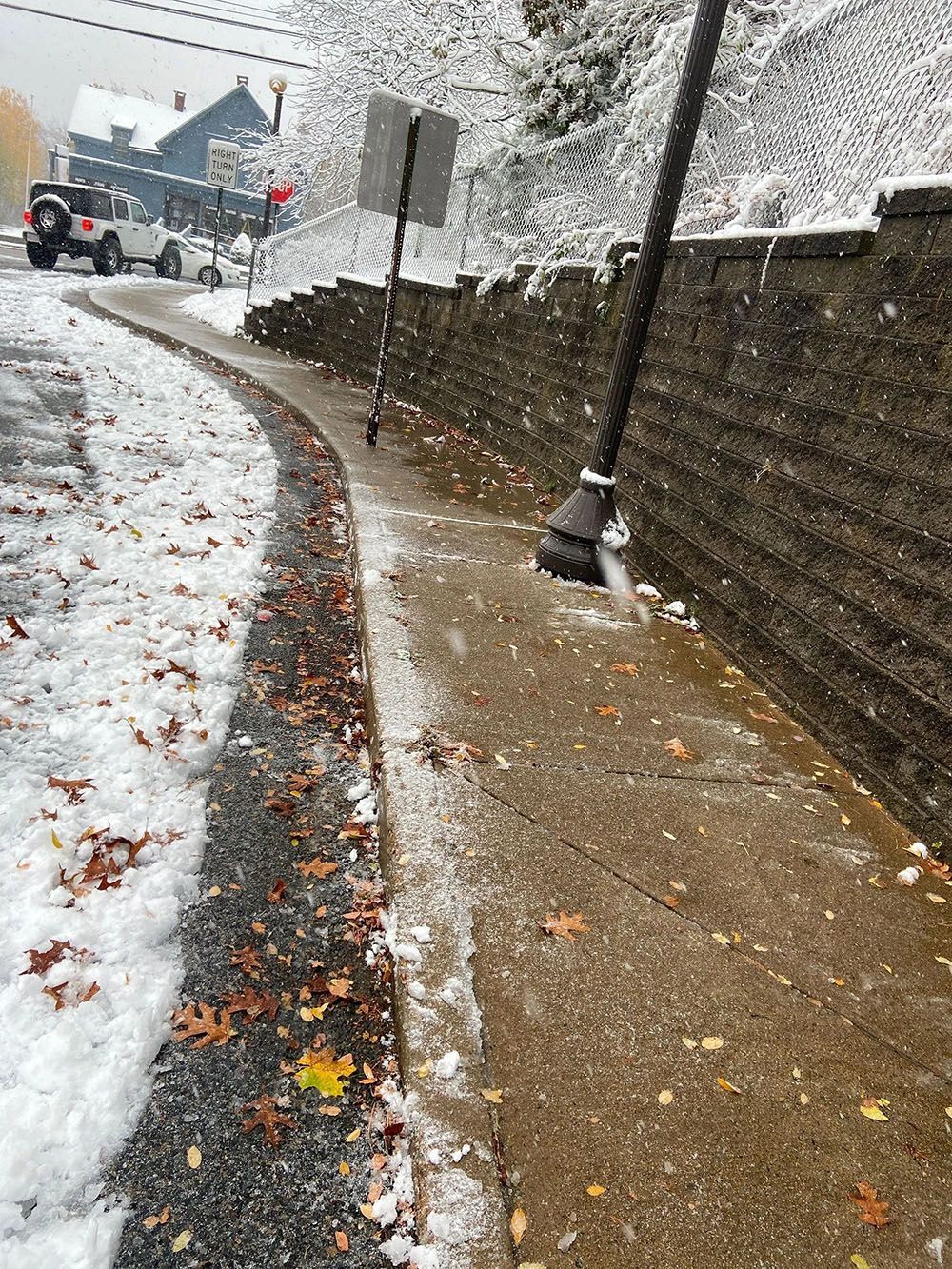 Snowy sidewalk beside a brick wall, with falling snow and a road curving left.