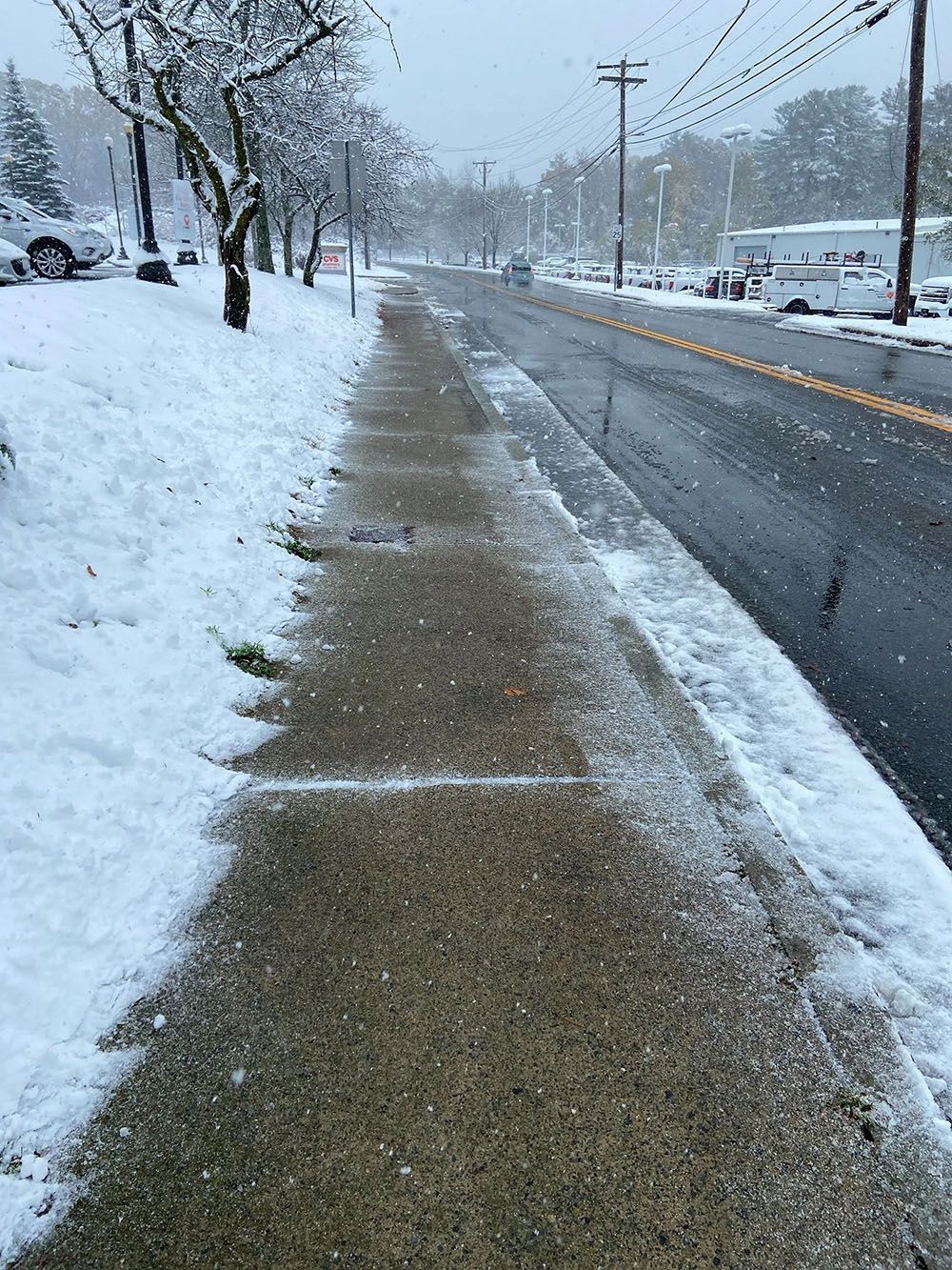 Snow-covered suburban sidewalk beside a wet road during a snowfall, with trees and houses in the background.