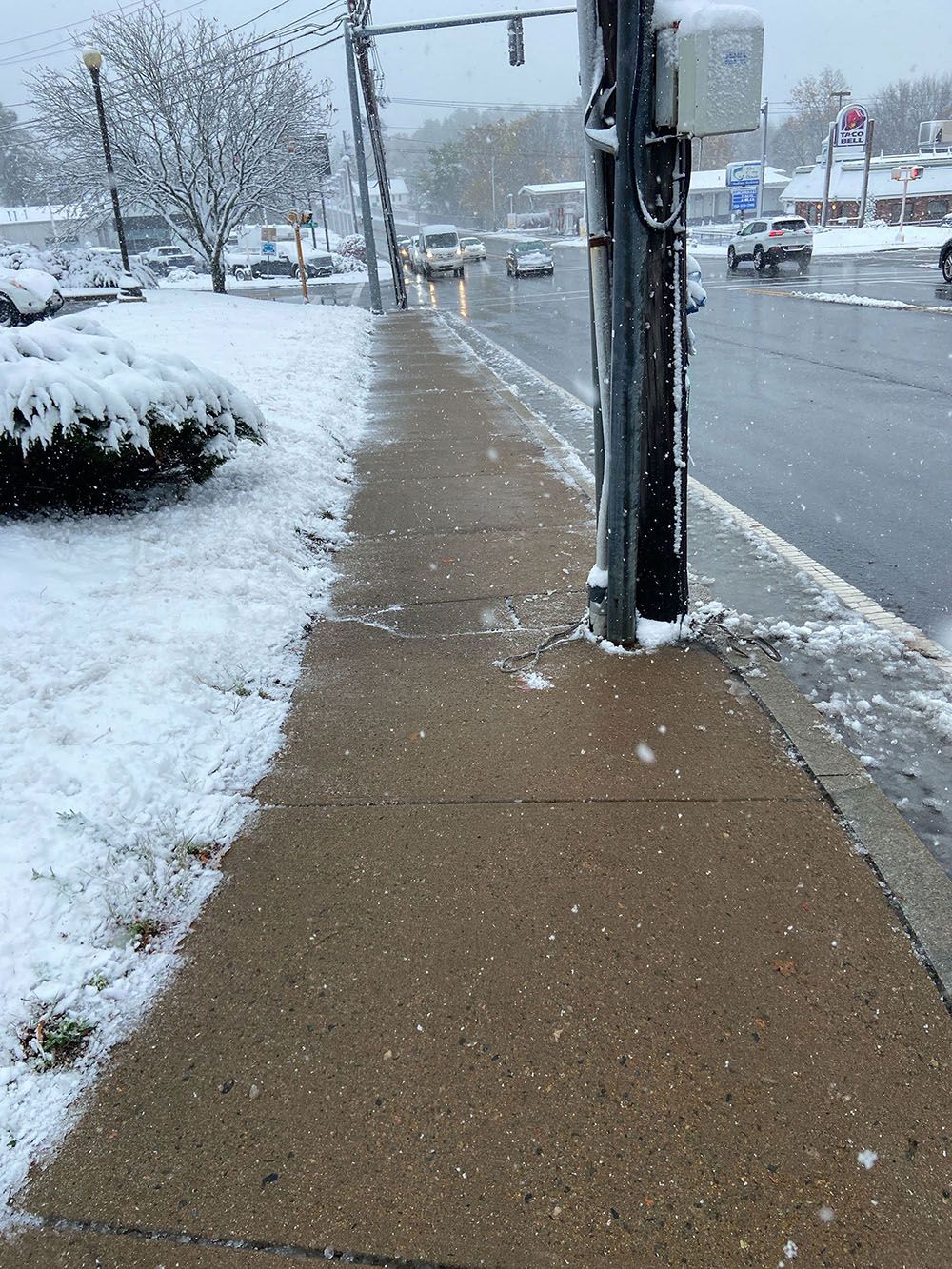 Snow-covered sidewalk beside a busy road in winter, with falling snow and a utility pole.