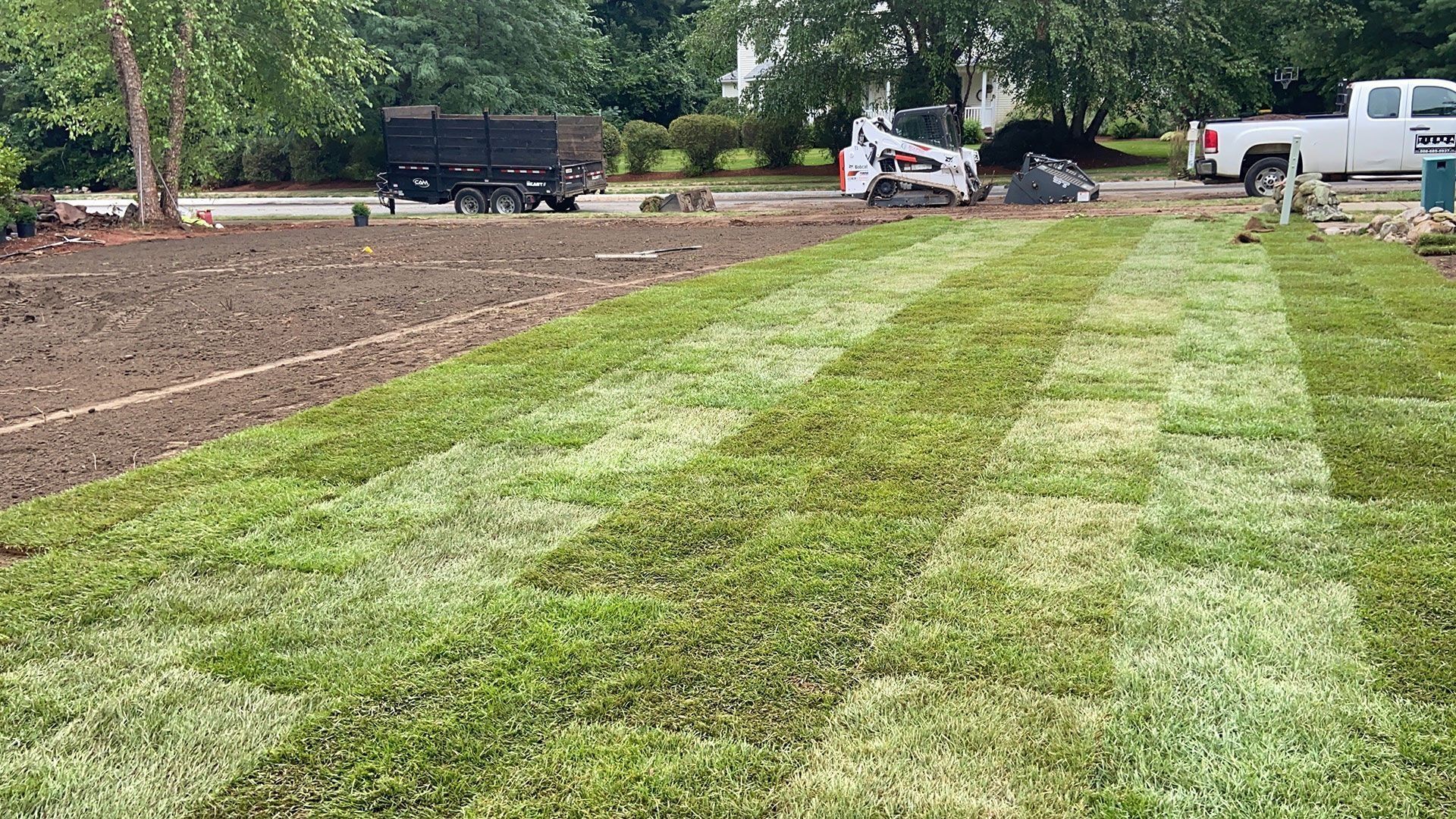 Freshly mowed lawn strips beside a dirt lot, with trucks and equipment in the background.