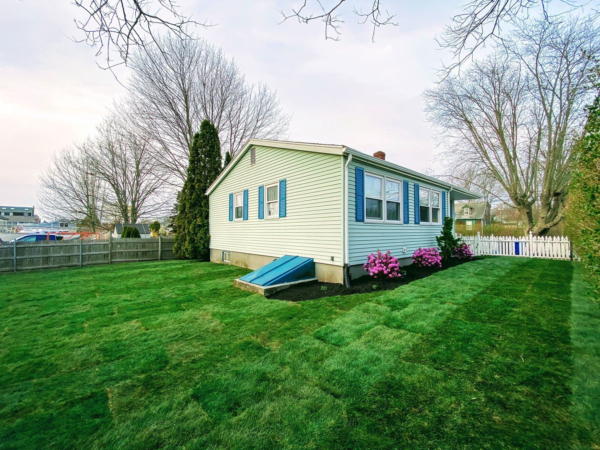 Single-story yellow house with blue shutters, pink flowers, and a large green lawn under a cloudy sky