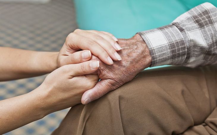 A man is talking to an elderly woman in a wheelchair while holding a cup of coffee.