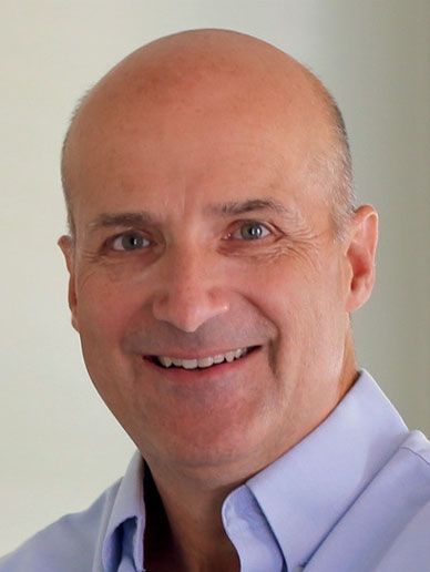 Bald man smiles, wearing a light blue collared shirt.